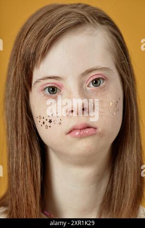 Portrait of girl with Down syndrome looking at camera, featuring freckles and stars on cheeks against smooth yellow background, expressing seriousness Stock Photo