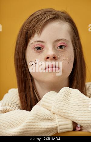 Portrait of young woman with Down syndrome having glitter on her cheeks and wearing white sweater, set against bright yellow background, resting head on hands Stock Photo