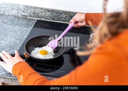 Over-the-shoulder shot of an unrecognizable woman preparing a fried egg in a pan on an induction cooker. Preparing a fried egg with its white and yolk Stock Photo