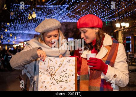 Girlfriends in coats and French berets with shopping bags enjoy cheerful Christmas shopping and a hot beverage at a festively decorated town square. Stock Photo