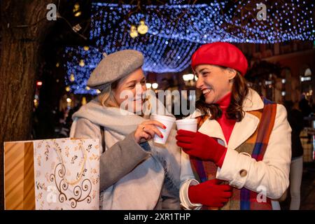 Girlfriends in coats and French berets with shopping bags enjoy cheerful Christmas shopping and a hot beverage at a festively decorated town square. Stock Photo