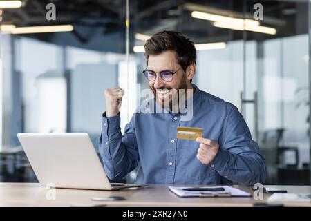 Excited man holding credit card and using laptop in modern office. Office worker expressing happiness, success, and accomplishment. Stock Photo