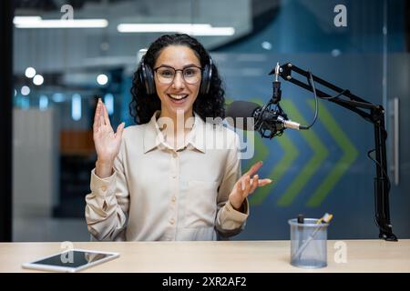 Smiling woman wearing headphones recording podcast with microphone in studio. Person gesturing hands during broadcast. Tablet phone on desk indicates use of technology. Stock Photo