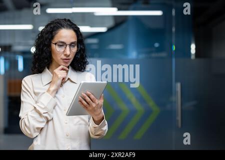 Focused businesswoman using tablet in modern office space. Analyzing data, making decisions, and working with technology. Professional attire highlighting determination and attention to detail. Stock Photo