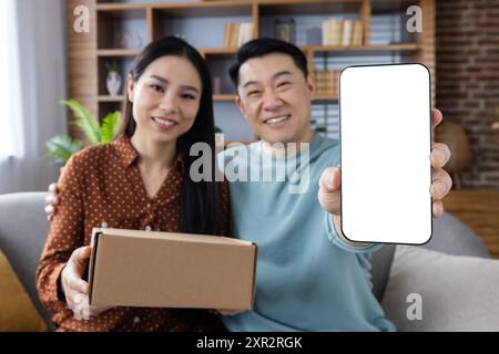 Asian couple sitting on sofa showing blank smartphone screen while holding cardboard box. Concept for online shopping, delivery service, technology, and modern lifestyle. Stock Photo