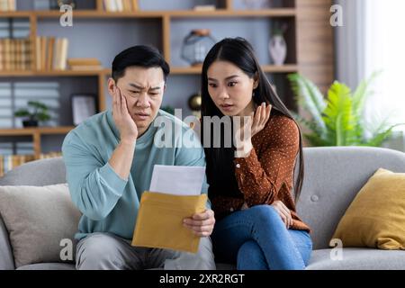 Asian couple sitting on couch looking stressed and worried while checking bills. Man and woman reviewing letters and financial documents with concern. Stock Photo