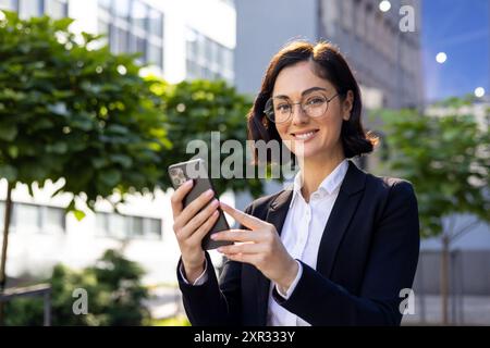 Portrait of a smiling young woman in a business suit standing in an office center and using a mobile phone, looking confidently at the camera. Stock Photo
