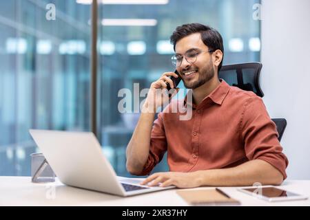 Smiling businessman sitting at desk, talking on phone while working on laptop. Office environment with professional working on project, multitasking, and staying connected. Stock Photo