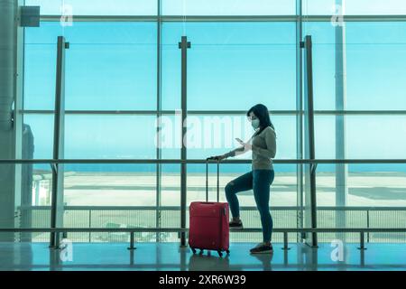 Woman with suitcase waiting in the terminal to board her flight and using her smartphone at the large window of the airport looking at the landing str Stock Photo