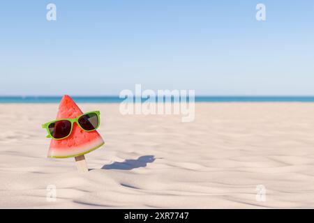 3d rendering of a whimsical watermelon slice on a stick sporting green sunglasses against a sunny beach backdrop with the ocean in the distance. Summe Stock Photo