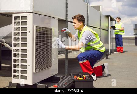 Technician installing and fixing air conditioner using screwdriver sitting on the roof of building. Stock Photo
