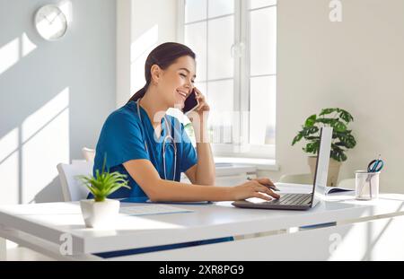 Female doctor in blue medical uniform sitting at the desk using laptop and talking by mobile phone. Stock Photo
