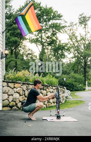 Man repairing a bicycle in a driveway with a Pride flag hanging nearby Stock Photo