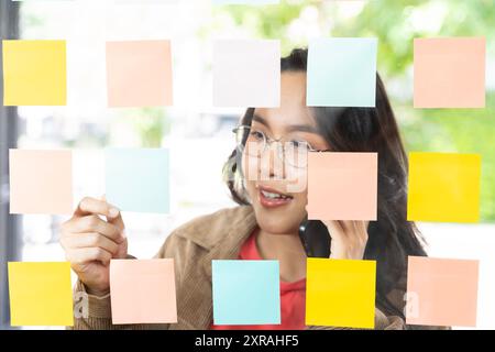 A businesswoman writes her goals on sticky notes and arranges them on a glass wall to map out her plan for success. Brainstorm her ideas and map out h Stock Photo