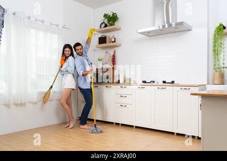 Beautiful loving young couple mopping floor in kitchen together, full length photo, copy space. Romantic cheerful lady hugging her smiling boyfriend w Stock Photo