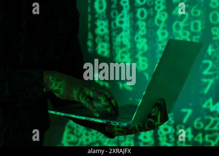 The hacker is wearing a hoodie and sunglasses, and their face is hidden in the shadows. The laptop screen is filled with code. A hacker is sitting in Stock Photo