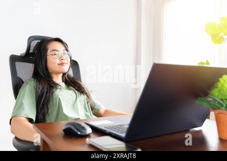 Young woman crossed hands behind head, enjoying break time at home. Peaceful carefree business woman resting at table with computer, looking aside, dr Stock Photo