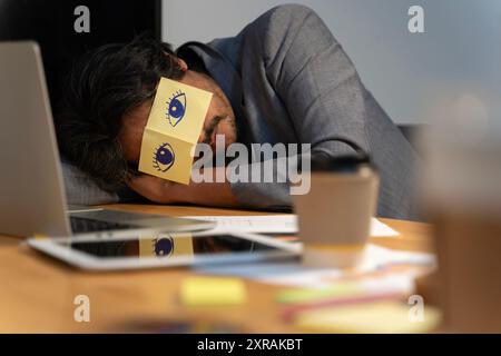 Cheerful adult man resting in office with sticky notes on the eyes. Sleeping man having stickers with eyes painted on face relaxing with hands behind Stock Photo