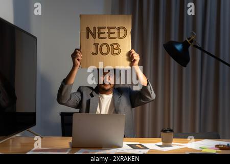 Job hunting. Man sitting at the table in the office, holding a piece of cardboard that says need job in his hands. Hire me concept. Businessman lookin Stock Photo