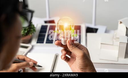 Close-up at a light bulb, An engineer sits holding a light bulb and model of wind turbine. with solar panels and model house on the table. To design u Stock Photo