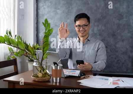Joyful Businessman Waving Hello Smiling To Camera Sitting In Modern Office. Entrepreneur Man Communicating Online Gesturing Hi Greeting At Workplace. Stock Photo