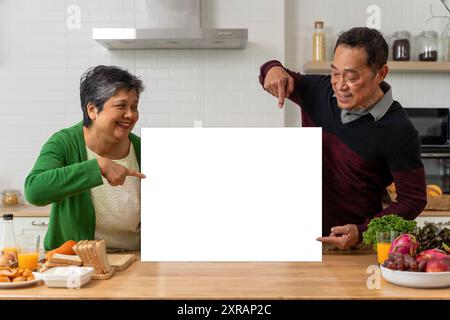 An elderly couple having breakfast together in the kitchen at home. Holding a blank white billboard placed on the table can contain advertising media, Stock Photo