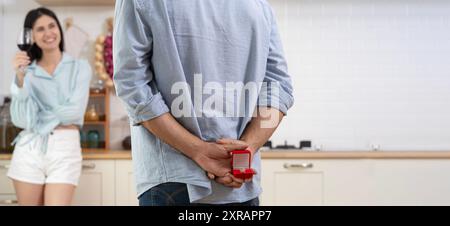 Multiethnic couple having romantic dinner in home kitchen. A man stands with a box with a wedding ring behind him, offering it to a woman who smiles i Stock Photo