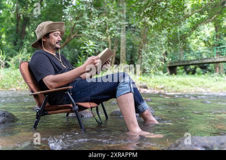 Reading or education concept. A happy handsome bearded Asian man sits alone in a chair near a stream in the countryside and enjoys the forest view whi Stock Photo