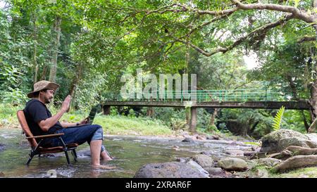 Attractive man relaxing in chair while video calling on laptop. Young handsome asian traveler relaxing and enjoying outdoor camping on wild mountain i Stock Photo