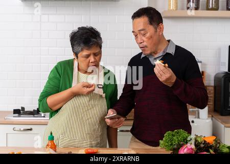 Senior couple on the phone looking for breakfast recipes in the kitchen at home. Retirement, hobbies chef skills on the internet on smartphones online Stock Photo
