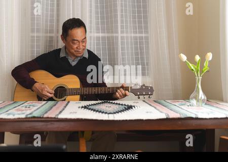 Senior man happy sitting playing acoustic guitar indoors as hobby after retirement. Elderly musician has fun singing a song with an instrument. Cheerf Stock Photo
