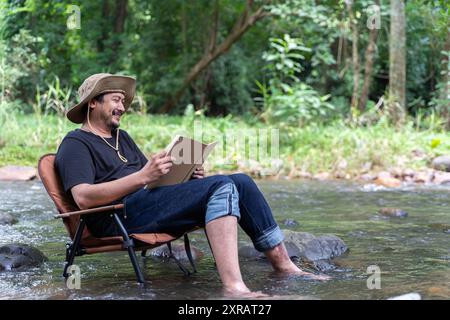 A happy handsome bearded Asian man sits alone in a chair near a stream in the countryside and enjoys the forest view while reading a book. Reading or Stock Photo