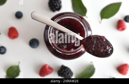 Detail of wild berry jam with spoon with jam on bowl and fruits in background on white table. Top view. Stock Photo