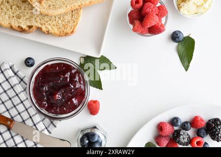 Breakfast with wild berry jam in a glass bowl on a white table with a bowl full of berries and slices of bread. Top view. Stock Photo