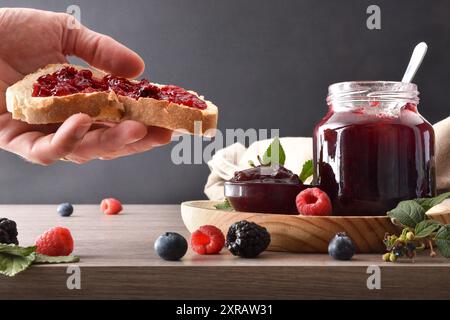 Hand showing slice of bread with berries jam and wooden kitchen bench with containers with jam and breakfast products and isolated dark background. Fr Stock Photo