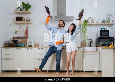 Happy young couple doing home chores, singing and having fun. Funny woman singing songs and dancing while cleaning kitchen at home, couple having fun Stock Photo