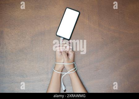 Woman hands trapped and wrapped on wrists with mobile phone cable as handcuffs in smart phone networking and communication technology addiction concep Stock Photo
