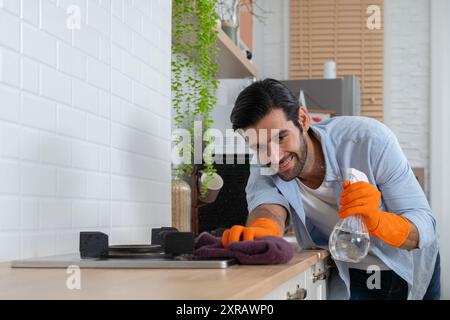 Young handsome man with apron and rubber gloves wiping wooden table in kitchen. Cleaning service concept. Wiping dining table with cloth and smiling. Stock Photo