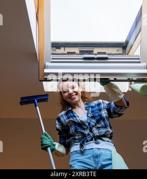 Happy young adult woman washing roof windows using squeegee. Stock Photo