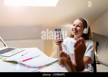 Joyful woman wearing a white t-shirt sits at the table with her feet up student room of university camp, using headphones and a cellphone. Stock Photo