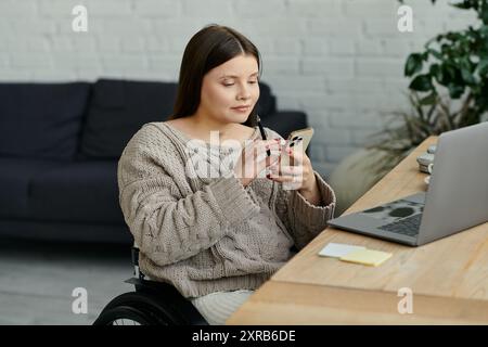 A young woman in a wheelchair sits at a table in her home, using her smartphone. Stock Photo