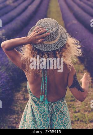 Back view of pretty babe in boho elegant blue dress walk in lavender fields wearing travel style hat and blonde curly nice long hair - concept or free woman in the outdoor nature adventure lifestyle Stock Photo