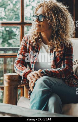 Hipster style pretty lady sitting at home on the sofa and looking outside thw windows a green forest woods. Cozy cabiness chalet female people relax lifestyle. Young adult woman enjoy leisure at home Stock Photo