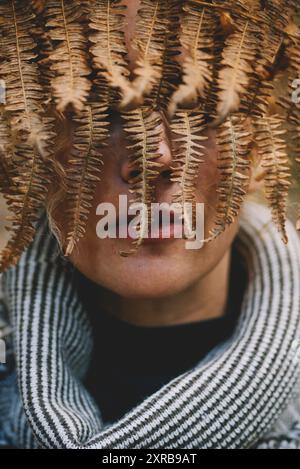 Concept of nature and environment save the world. Close-up portrait of woman hidden his face with dry autumn leaf in front of her face. Park and green forest in background Stock Photo