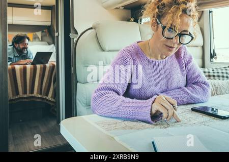 People man and woman in van life day inside camper van. Woman using phone gps map to plan next destination and man working on computer in background. Couple in travel lifestyle with vehicle on caravan Stock Photo