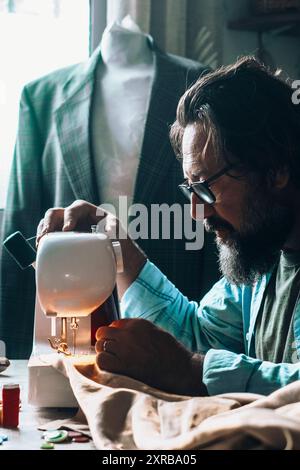 One man in home workplace use sewing machine to work on vintage garment alone. Male people working as a tailor indoor. Alternative business store concept lifestyle. Young adult in garment production Stock Photo