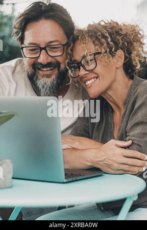 One couple middle age enjoy and have fun together using laptop sitting outdoor at the table in the garden at home. Man and woman in love and friendship doing video web call together smiling. Happiness Stock Photo