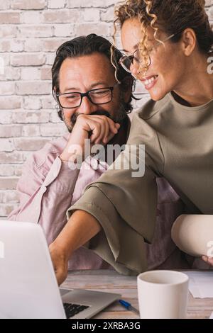 Happy couple of adult worker smile and enjoy job together. Woman pointing at computer display and man laugh. Adult people with laptop and papers. Collegues at work in home office on notebook pc Stock Photo