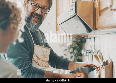 Real couple cooking together at home in the kitchen. One man smile to a woman and using pots preparing lunch. Husband and wife living in apartment. Mature people in indoor leisure activity happiness Stock Photo