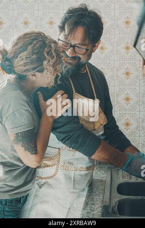One man helping wife in housework job washing the dishes in the kitchen. Woman appreciate husband help in housekeeping leisure activity. Couple smile and hug with love inside home cleaning together Stock Photo
